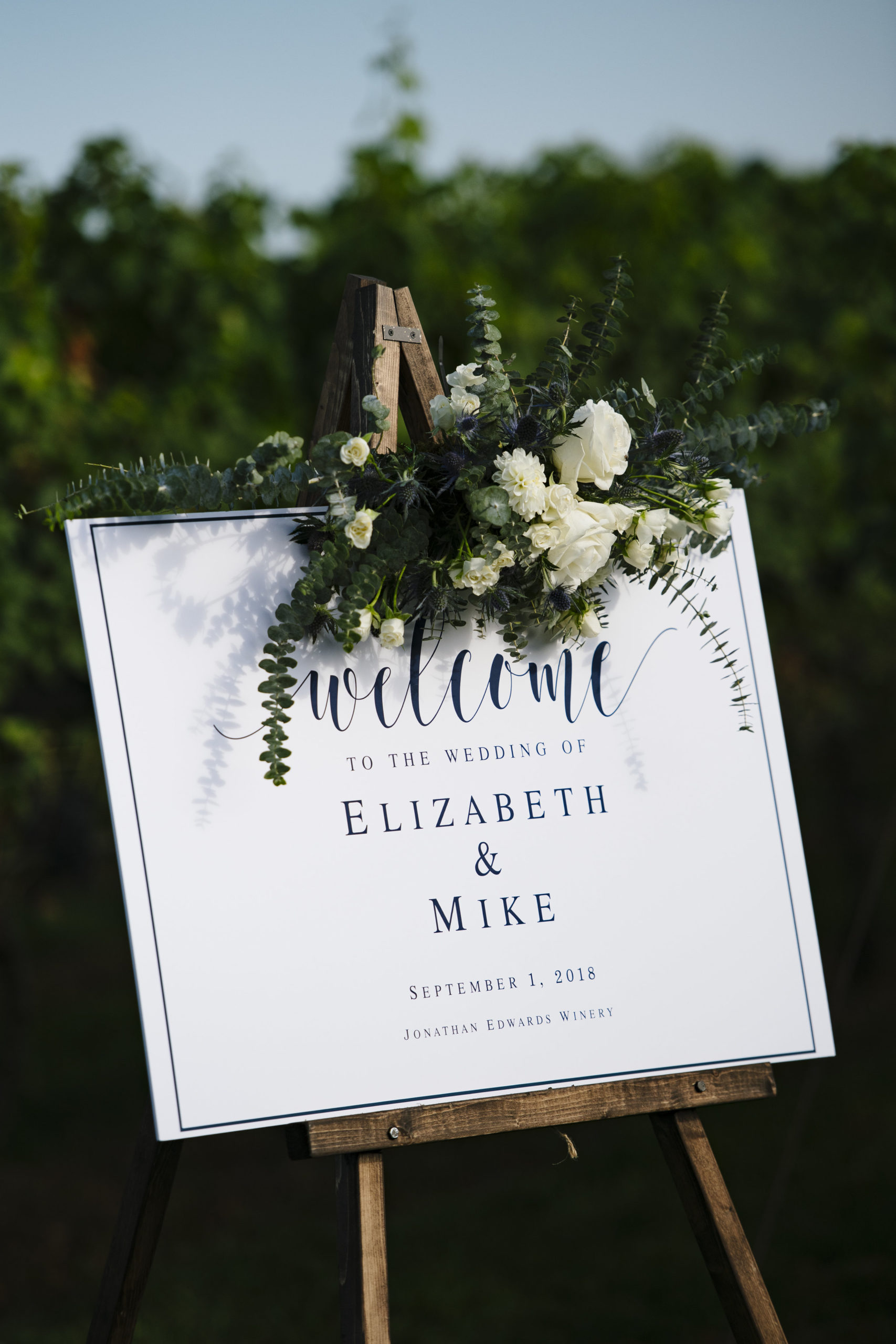 White and navy blue welcome sign on an easel with florals at the top at Jonathan Edward Winery in Connecticut - Pearl Weddings &amp; Events