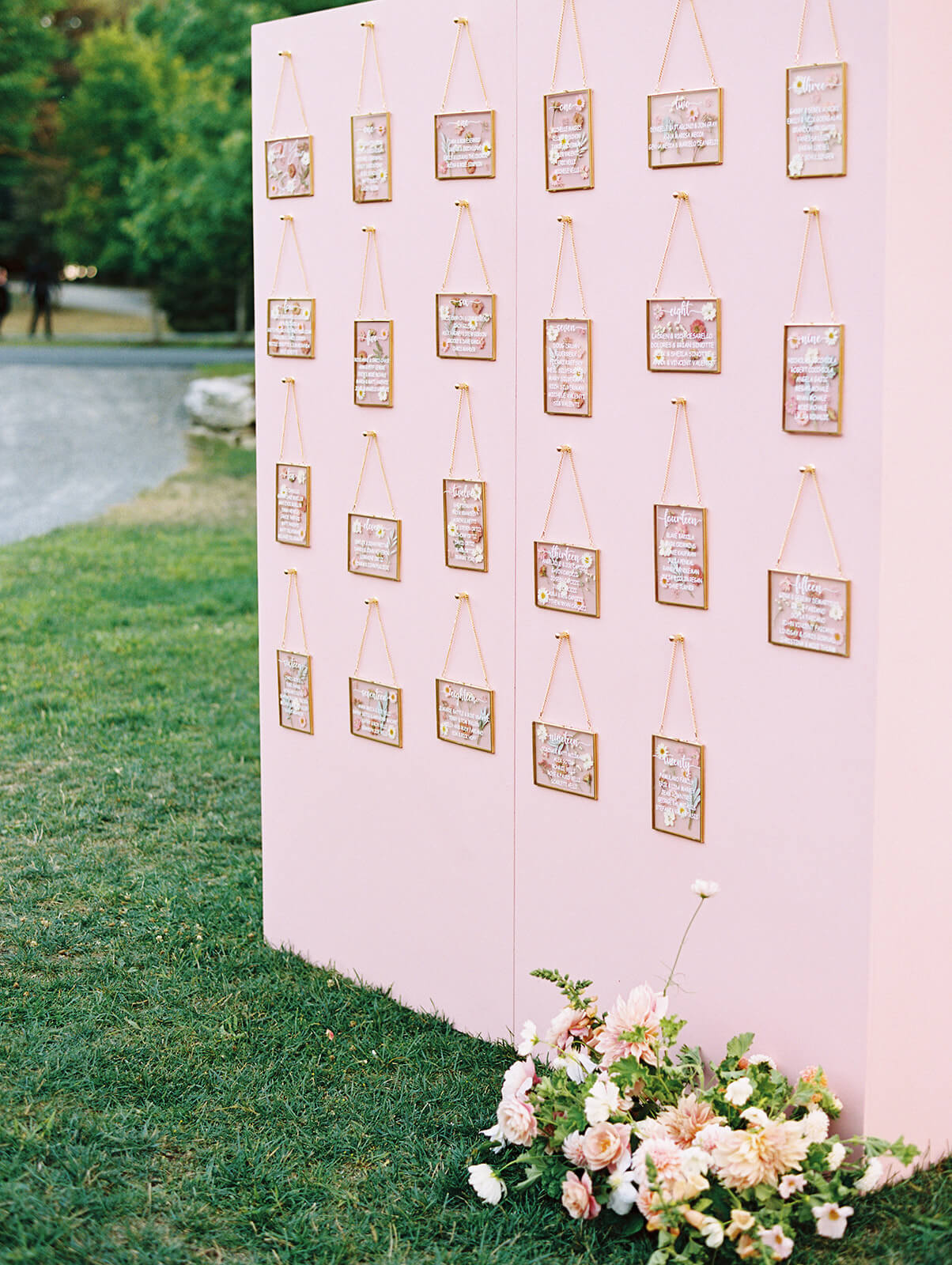 Hanging on a pink wall, is clear frames with pressed dried flowers for a unique seating chart